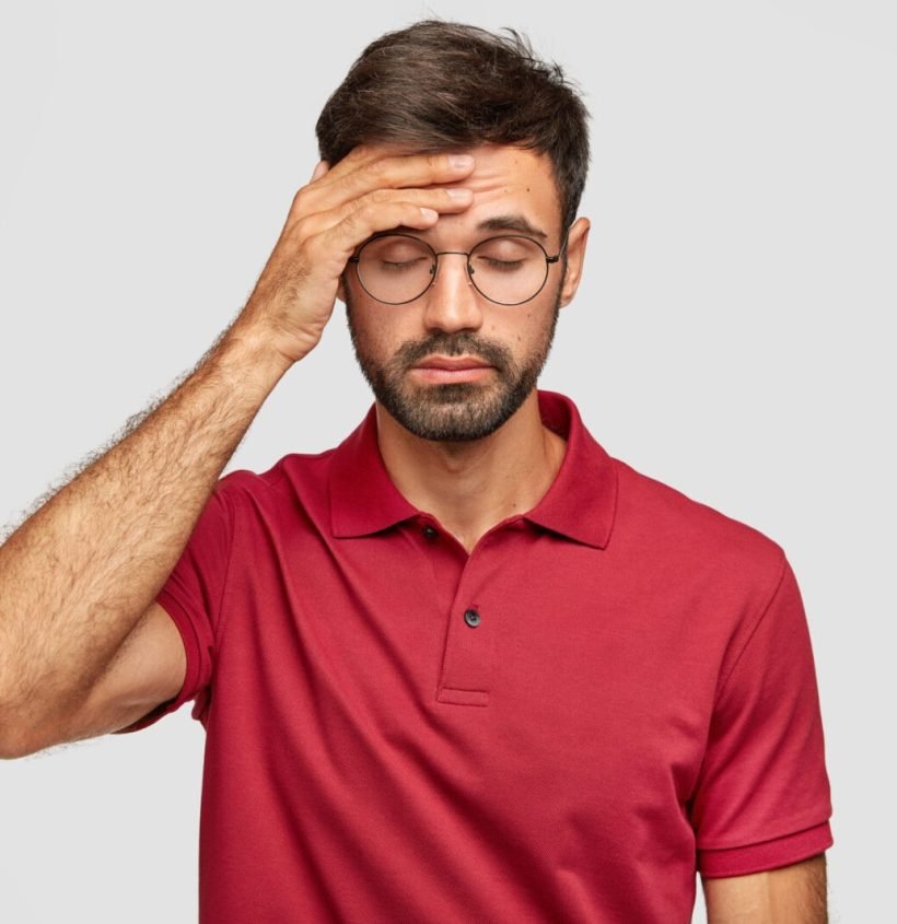 Overworked Cuacasian male feels terrible headache after sleepless night, keeps hand on forehead, shut eyes, dressed in casual red t shirt, stands against white background. People and tiredness